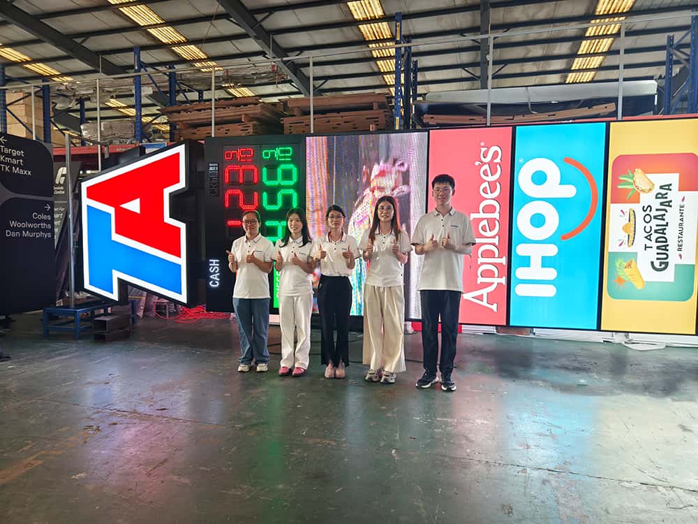 Four factory workers standing in front of a giant multi-tenant highway pylon sign for a TA truck stop, demonstrating its massive scale.
